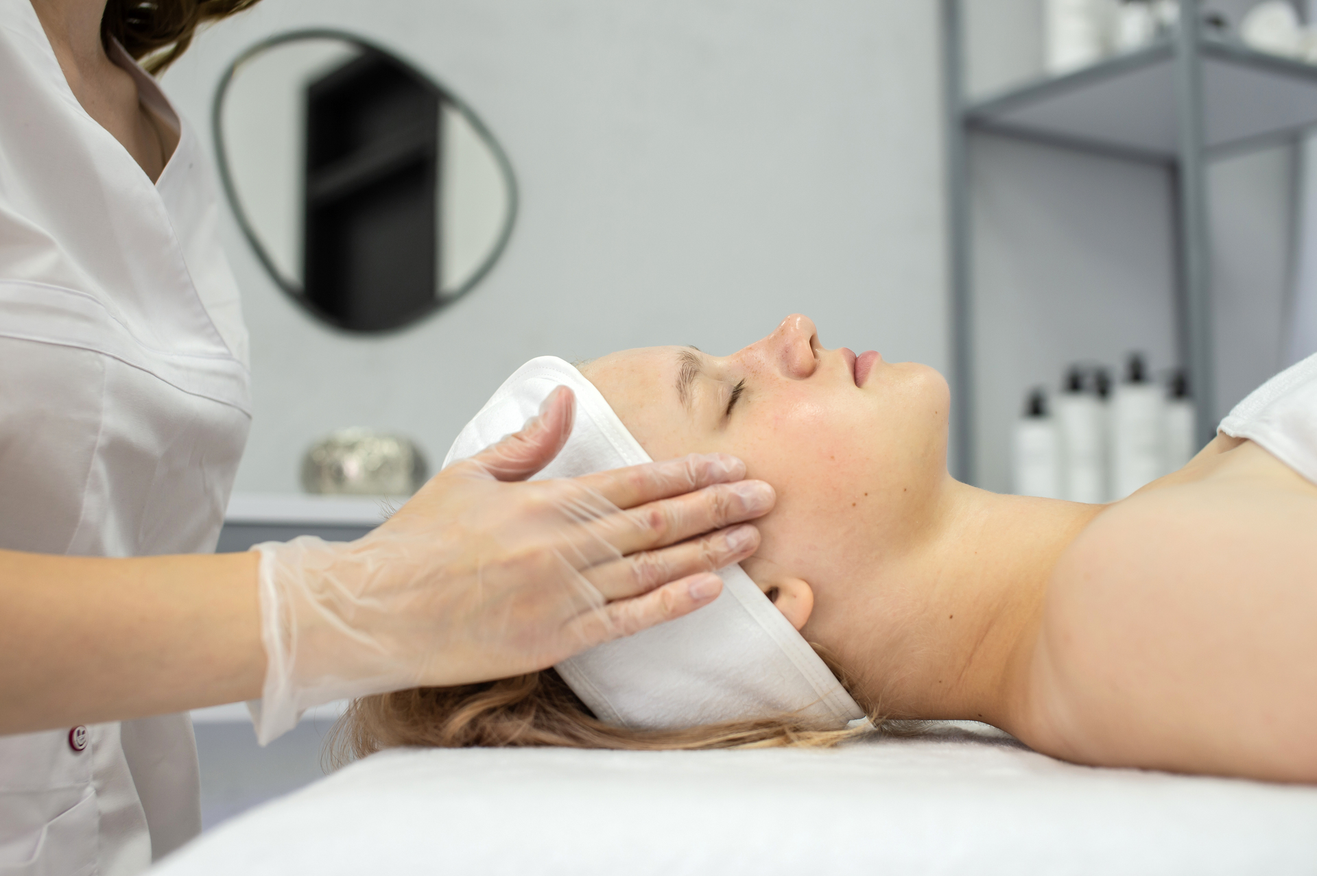 Woman receiving a facial treatment while lying down, with a therapist gently touching her face wearing gloves.