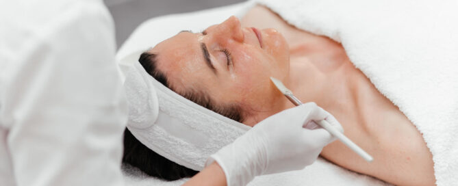A woman receiving a facial treatment while lying down with a white headband, as an aesthetician wearing white gloves applies product to his face with a brush.