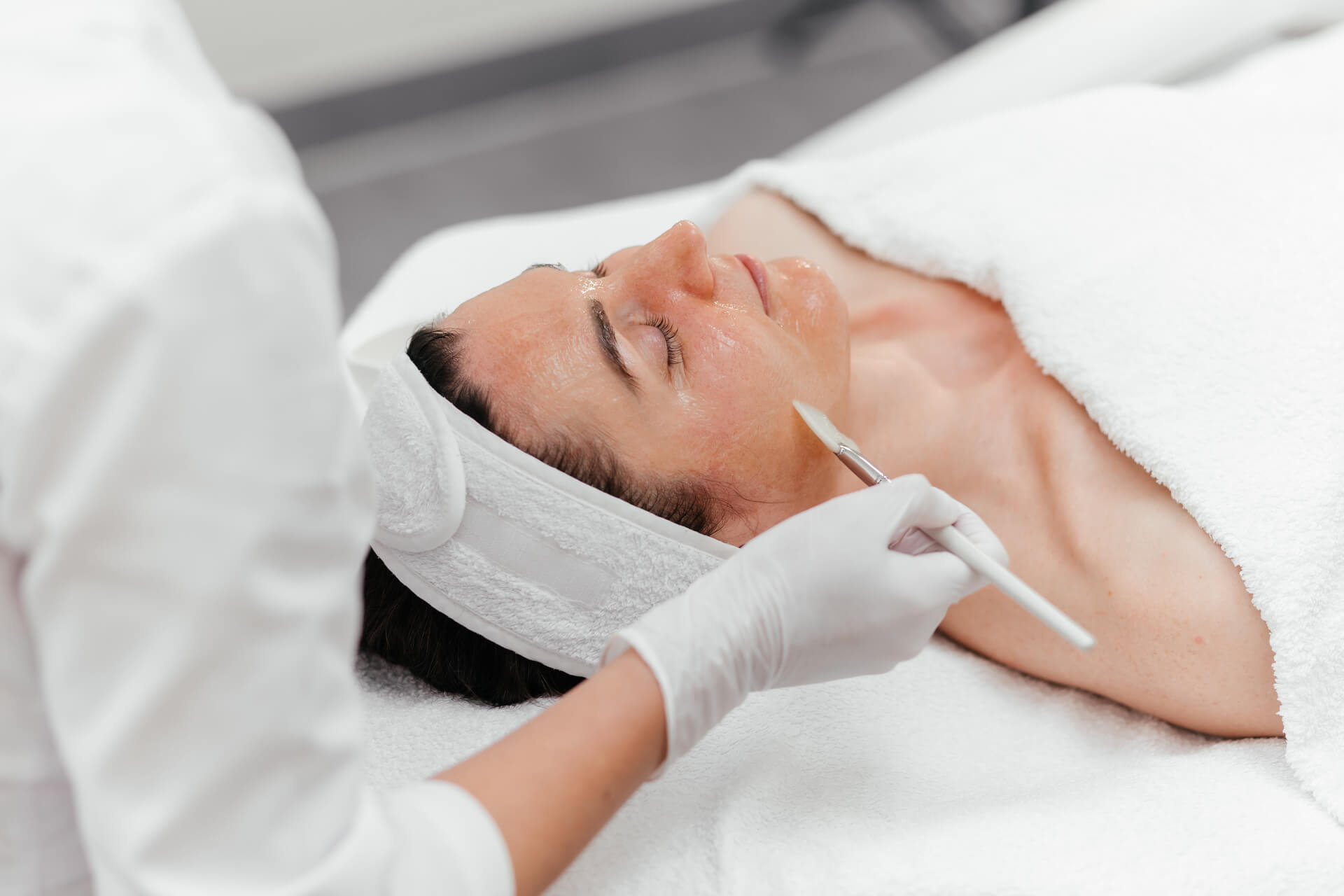 A woman receiving a facial treatment while lying down with a white headband, as an aesthetician wearing white gloves applies product to his face with a brush.